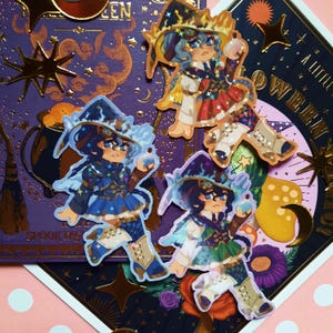 May include: Three witch stickers with glitter accents. Each witch is wearing a different colored outfit and hat. The witches are holding a potion bottle and a wand. The stickers are on a purple and gold background with stars.