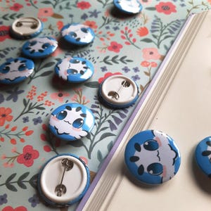 May include: A collection of round, blue and white enamel pins with a cartoon character design. The pins are scattered on a floral patterned surface, with some pins showing the pin back. The pins are approximately 1 inch in diameter.