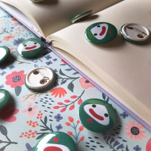 May include: Green button pins with a smiling face design, scattered on a floral patterned surface next to an open book. The pins have a white face with a red smile and are surrounded by a green border. Some pins are face up, and some are face down.