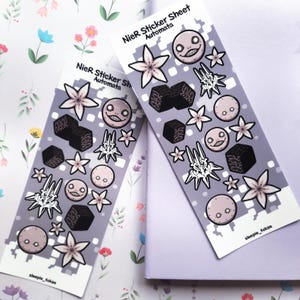May include: Two NieR:Automata sticker sheets with various character designs. The stickers feature floral, geometric, and character illustrations in shades of gray, black, and pink. The text "NieR Sticker Sheet Automata" is printed on the top of each sheet.