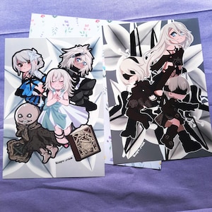 May include: Two art prints featuring anime-style characters. One print showcases a group of characters in black and white, while the other features a group of characters in various outfits. The prints are on a purple surface.