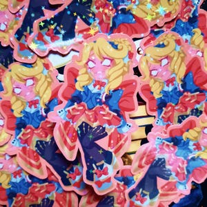 以下が含まれることがあります： 金髪で青い服と赤いリボンを着た漫画のキャラクターが描かれた、ホログラフィック加工が施されたカラフルなステッカーのセット。キャラクターは驚いた表情をしていて、魔法の杖を持っている。