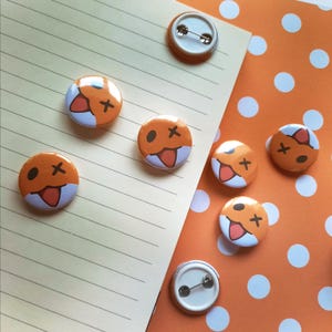 May include: Six orange and white button pins with a cartoon fox face on them. The fox has a big smile and a tongue sticking out. The buttons are on a white and orange polka dot background.