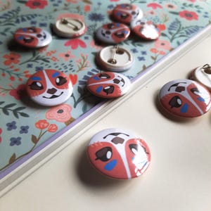 May include: A collection of round pinback buttons with a dog face design. The buttons feature a white background with a red and blue dog face illustration. Some buttons are face up, others show the pin back.