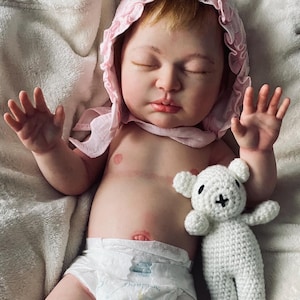 Puede incluir: Una mu&ntilde;eca beb&eacute; realista con un gorro rosa y un pa&ntilde;al, acostada sobre una manta blanca. Un peque&ntilde;o oso de peluche blanco tejido a ganchillo descansa junto a la mu&ntilde;eca. La mu&ntilde;eca tiene los ojos cerrados y piel clara.