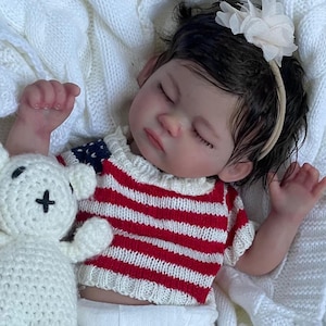 Può includere: Una bambola realistica con un maglione a righe rosse e bianche e un pannolino. La bambola è sdraiata su una coperta bianca lavorata a maglia accanto a un orsacchiotto bianco all'uncinetto. La bambola ha i capelli scuri e indossa una fascia con un fiore bianco.