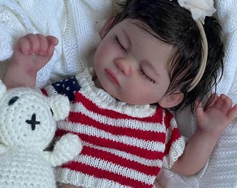 Puede incluir: Una muñeca bebé realista con un suéter a rayas rojas y blancas y un pañal. La muñeca está acostada sobre una manta blanca tejida junto a un oso de peluche blanco tejido a ganchillo. La muñeca tiene cabello oscuro y lleva una diadema con una flor blanca.