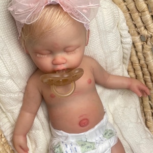 May include: A realistic newborn baby doll with blonde hair, wearing a large pink organza bow headband and a white diaper. The doll has closed eyes, a light skin tone, and a translucent pacifier in its mouth, resting in a woven basket.