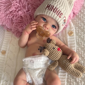 Può includere: Una bambola con un cappello di lana color crema con "MINNIE" ricamato sopra e un ciuccio. La bambola tiene in mano un piccolo giocattolo a forma di renna all'uncinetto. La bambola è sdraiata su una coperta di lana bianca con un cuscino rosa soffice.