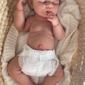 Puede incluir: Una mu&ntilde;eca reborn realista con cabello rizado casta&ntilde;o claro y ojos cerrados, acostada sobre una manta de punto blanca dentro de una cesta de mimbre tejida. La mu&ntilde;eca tiene un tono de piel claro, un ombligo detallado y lleva un pa&ntilde;al desechable blanco.