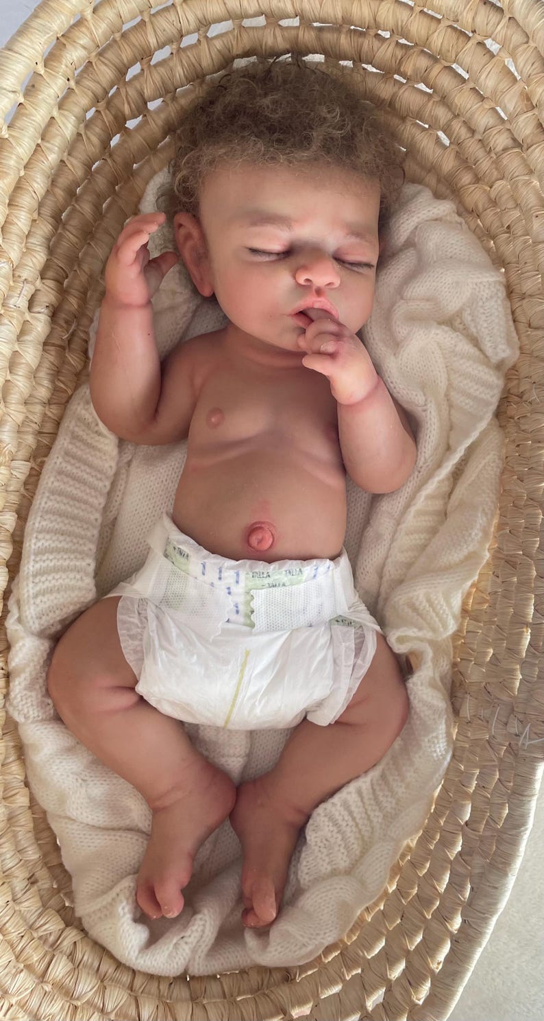 Puede incluir: Una mu&ntilde;eca reborn realista con cabello rizado casta&ntilde;o claro y ojos cerrados, acostada sobre una manta de punto blanca dentro de una cesta de mimbre tejida. La mu&ntilde;eca tiene un tono de piel claro, un ombligo detallado y lleva un pa&ntilde;al desechable blanco.