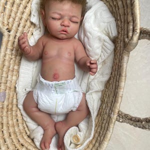 Puede incluir: Una mu&ntilde;eca reci&eacute;n nacida realista con cabello casta&ntilde;o claro y ojos cerrados, que lleva un pa&ntilde;al desechable blanco. La mu&ntilde;eca descansa dentro de una cesta de mimbre tejida forrada con una manta de punto trenzado blanco suave con un bot&oacute;n visible.