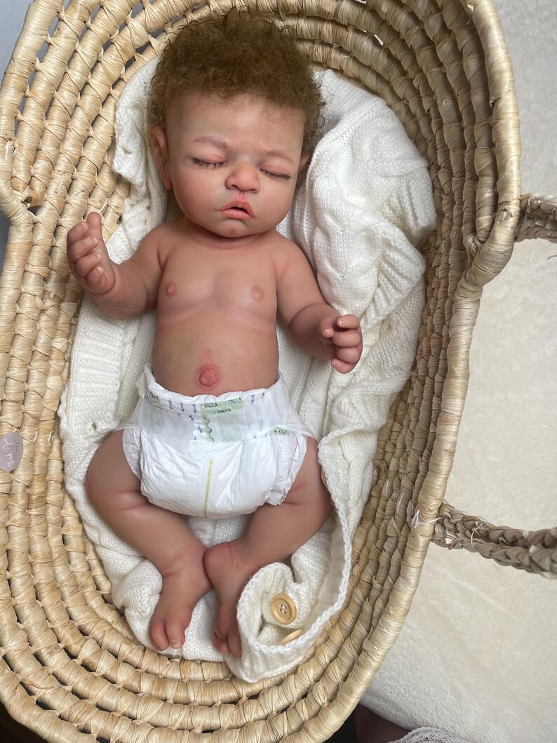 Puede incluir: Una mu&ntilde;eca reci&eacute;n nacida realista con cabello casta&ntilde;o claro y ojos cerrados, que lleva un pa&ntilde;al desechable blanco. La mu&ntilde;eca descansa dentro de una cesta de mimbre tejida forrada con una manta de punto trenzado blanco suave con un bot&oacute;n visible.