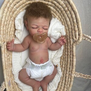 Puede incluir: Una mu&ntilde;eca reci&eacute;n nacida realista con cabello rizado casta&ntilde;o claro y ojos cerrados, que lleva un pa&ntilde;al blanco y un chupete transparente. La mu&ntilde;eca est&aacute; acostada sobre una manta de punto blanca dentro de una cesta de mimbre tejida.