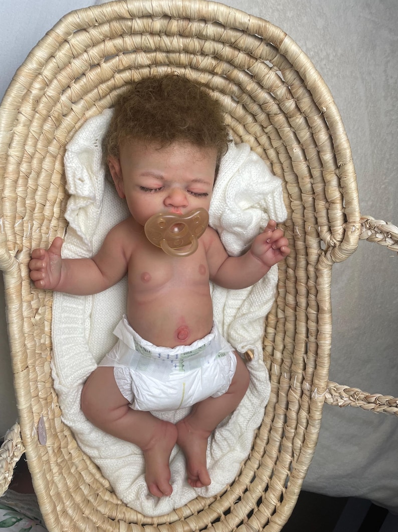Puede incluir: Una mu&ntilde;eca reci&eacute;n nacida realista con cabello rizado casta&ntilde;o claro y ojos cerrados, que lleva un pa&ntilde;al blanco y un chupete transparente. La mu&ntilde;eca est&aacute; acostada sobre una manta de punto blanca dentro de una cesta de mimbre tejida.