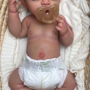Puede incluir: Una mu&ntilde;eca reci&eacute;n nacida realista con piel clara, cabello casta&ntilde;o rizado y ojos cerrados, que lleva un pa&ntilde;al desechable blanco y un chupete transl&uacute;cido. La mu&ntilde;eca est&aacute; acostada sobre una manta de punto blanca dentro de una cesta tejida.
