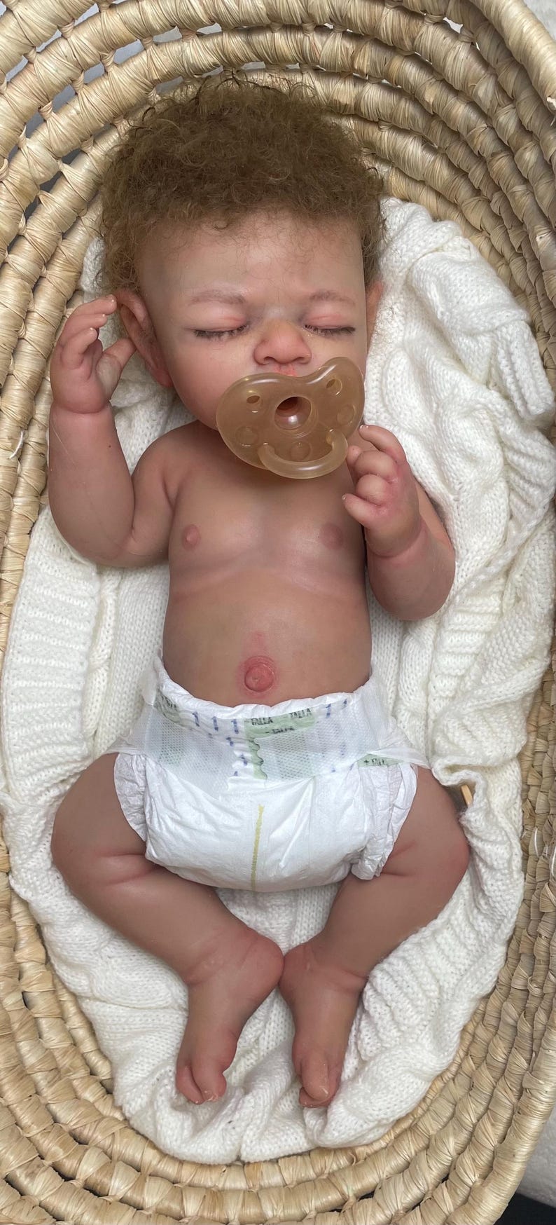 Puede incluir: Una mu&ntilde;eca reci&eacute;n nacida realista con piel clara, cabello casta&ntilde;o rizado y ojos cerrados, que lleva un pa&ntilde;al desechable blanco y un chupete transl&uacute;cido. La mu&ntilde;eca est&aacute; acostada sobre una manta de punto blanca dentro de una cesta tejida.