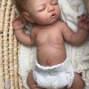 Puede incluir: Una mu&ntilde;eca reborn realista de piel clara, cabello casta&ntilde;o rizado y ojos cerrados, acostada en una cesta tejida. La mu&ntilde;eca lleva un pa&ntilde;al desechable blanco y est&aacute; colocada sobre una manta de punto blanca, mostrando detalles realistas.