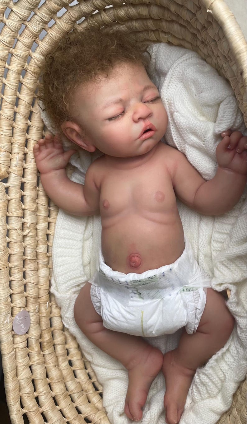 Puede incluir: Una mu&ntilde;eca reborn realista de piel clara, cabello casta&ntilde;o rizado y ojos cerrados, acostada en una cesta tejida. La mu&ntilde;eca lleva un pa&ntilde;al desechable blanco y est&aacute; colocada sobre una manta de punto blanca, mostrando detalles realistas.