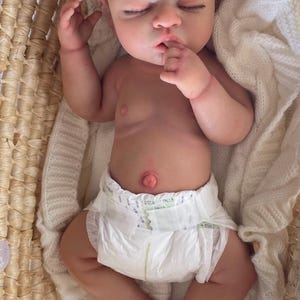 Puede incluir: Una mu&ntilde;eca realista de reci&eacute;n nacido con cabello rizado casta&ntilde;o claro y ojos cerrados, acostada boca arriba. La mu&ntilde;eca tiene una tez clara, un ombligo prominente y lleva puesto un pa&ntilde;al desechable blanco con una mano cerca de la boca.