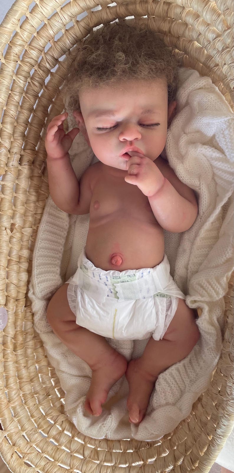 Puede incluir: Una mu&ntilde;eca realista de reci&eacute;n nacido con cabello rizado casta&ntilde;o claro y ojos cerrados, acostada boca arriba. La mu&ntilde;eca tiene una tez clara, un ombligo prominente y lleva puesto un pa&ntilde;al desechable blanco con una mano cerca de la boca.