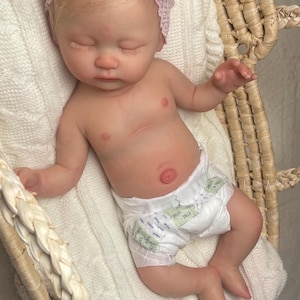 May include: A realistic newborn doll with light skin and blonde hair, wearing a white diaper and a pale pink lace headband with a large bow. The doll has closed eyes and is lying in a woven basket lined with a soft, white, textured fabric.