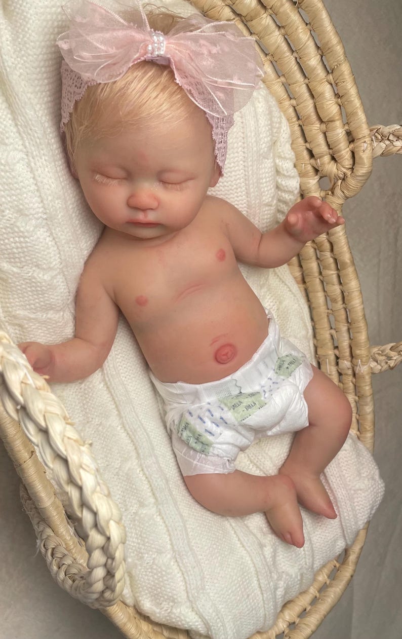 May include: A realistic newborn doll with light skin and blonde hair, wearing a white diaper and a pale pink lace headband with a large bow. The doll has closed eyes and is lying in a woven basket lined with a soft, white, textured fabric.