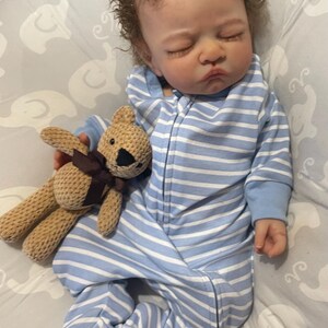 May include: A lifelike baby doll dressed in a blue and white striped onesie with a hood. The doll holds a small brown teddy bear with a dark brown ribbon. The baby has curly brown hair and rests on a grey patterned surface.