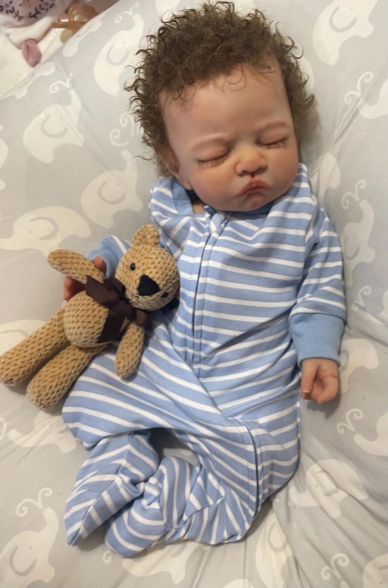 May include: A lifelike baby doll dressed in a blue and white striped onesie with a hood. The doll holds a small brown teddy bear with a dark brown ribbon. The baby has curly brown hair and rests on a grey patterned surface.