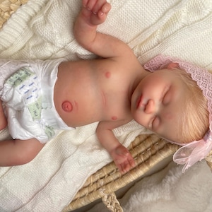 May include: A realistic newborn baby doll with light skin and blonde hair, wearing a white disposable diaper and a light pink lace headband with a matching bow. The doll is lying down with eyes closed, featuring detailed skin texture and a navel.