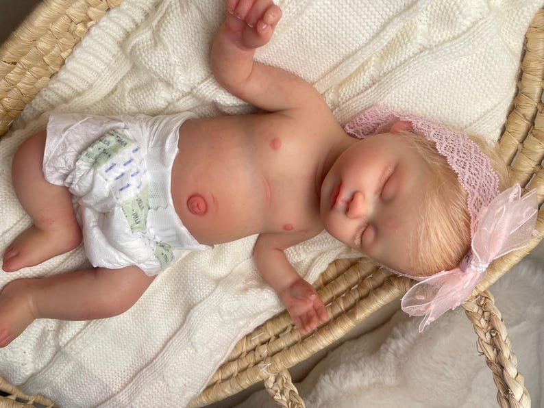 May include: A realistic newborn baby doll with light skin and blonde hair, wearing a white disposable diaper and a light pink lace headband with a matching bow. The doll is lying down with eyes closed, featuring detailed skin texture and a navel.