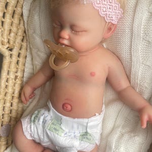 May include: A realistic newborn baby doll with light skin and blonde hair, wearing a pink lace headband with a bow and a white diaper. The doll has closed eyes, a light-colored pacifier in its mouth, and is resting on a white knitted blanket.
