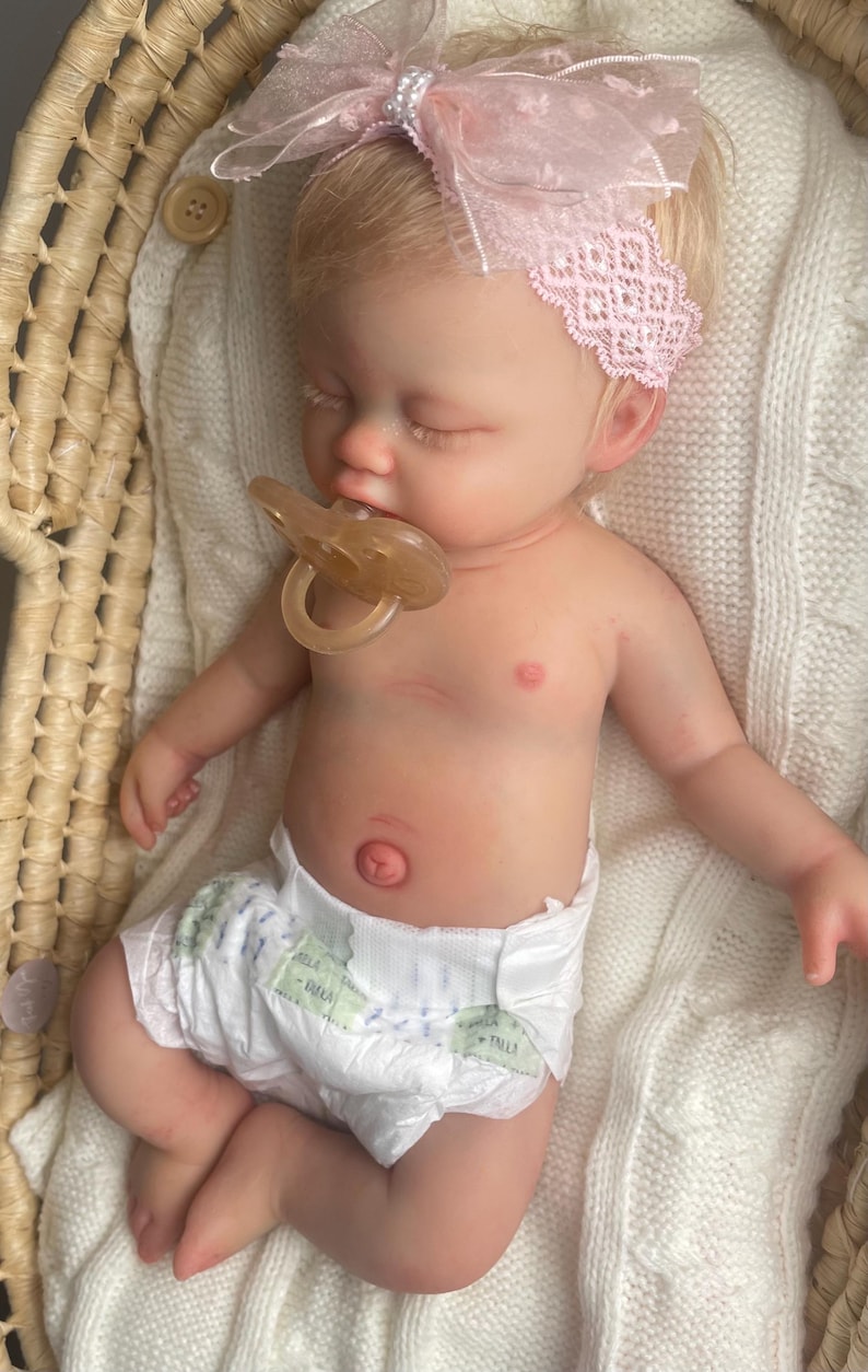 May include: A realistic newborn baby doll with light skin and blonde hair, wearing a pink lace headband with a bow and a white diaper. The doll has closed eyes, a light-colored pacifier in its mouth, and is resting on a white knitted blanket.