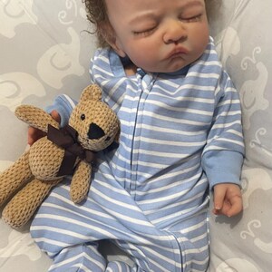 May include: A realistic baby doll dressed in a blue and white striped onesie with a zip. The doll has brown, curly hair and is holding a small, tan teddy bear with a brown ribbon. The doll is lying on a grey patterned blanket.