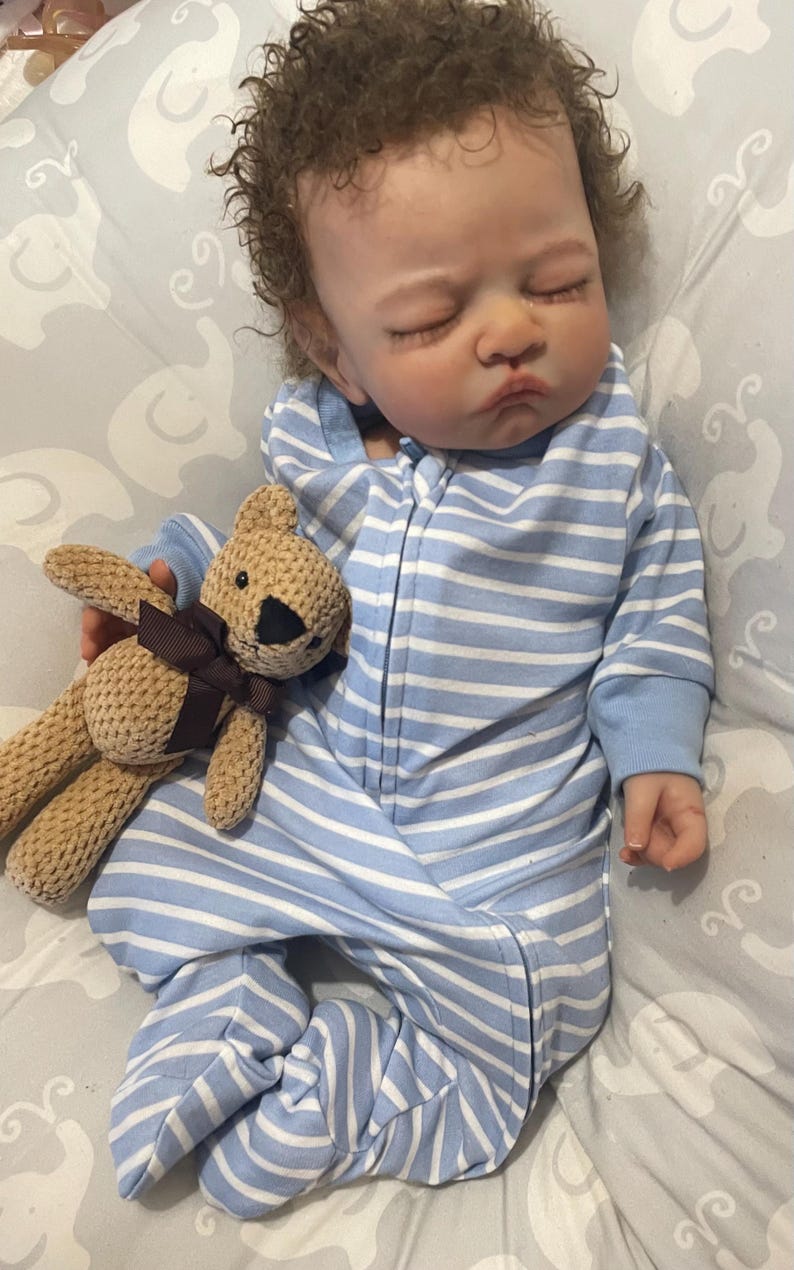 May include: A realistic baby doll dressed in a blue and white striped onesie with a zip. The doll has brown, curly hair and is holding a small, tan teddy bear with a brown ribbon. The doll is lying on a grey patterned blanket.
