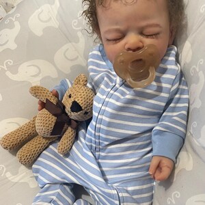 May include: A lifelike baby doll in a blue and white striped sleepsuit, with a dummy in its mouth. The doll clutches a small, beige teddy bear with a brown bow. The baby has brown, curly hair and rests on a grey patterned blanket.