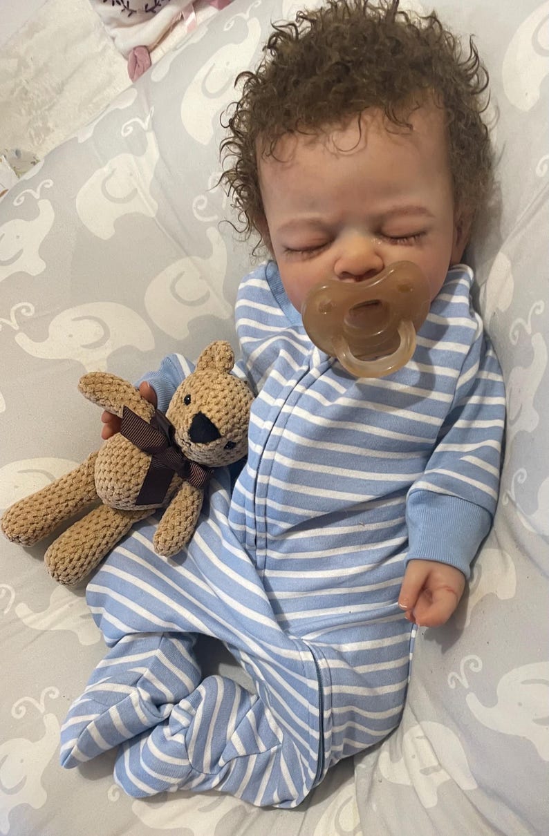 May include: A lifelike baby doll in a blue and white striped sleepsuit, with a dummy in its mouth. The doll clutches a small, beige teddy bear with a brown bow. The baby has brown, curly hair and rests on a grey patterned blanket.