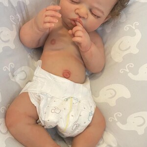 May include: A realistic baby doll with brown, curly hair and closed eyes, wearing a white nappy. The doll is lying on a grey patterned surface. The doll's skin has a realistic tone.