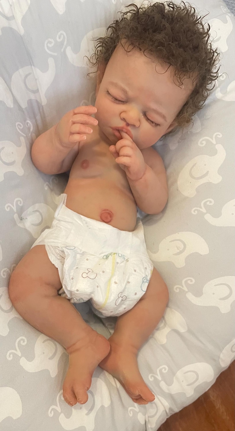 May include: A realistic baby doll with brown, curly hair and closed eyes, wearing a white nappy. The doll is lying on a grey patterned surface. The doll's skin has a realistic tone.