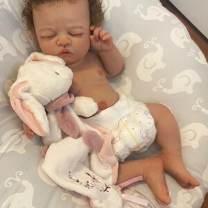 May include: A realistic baby doll with brown curly hair, wearing a nappy, and resting on a grey elephant-patterned pillow. The doll is holding a white and pink plush bunny. The bunny has pink ears and a pink scarf.