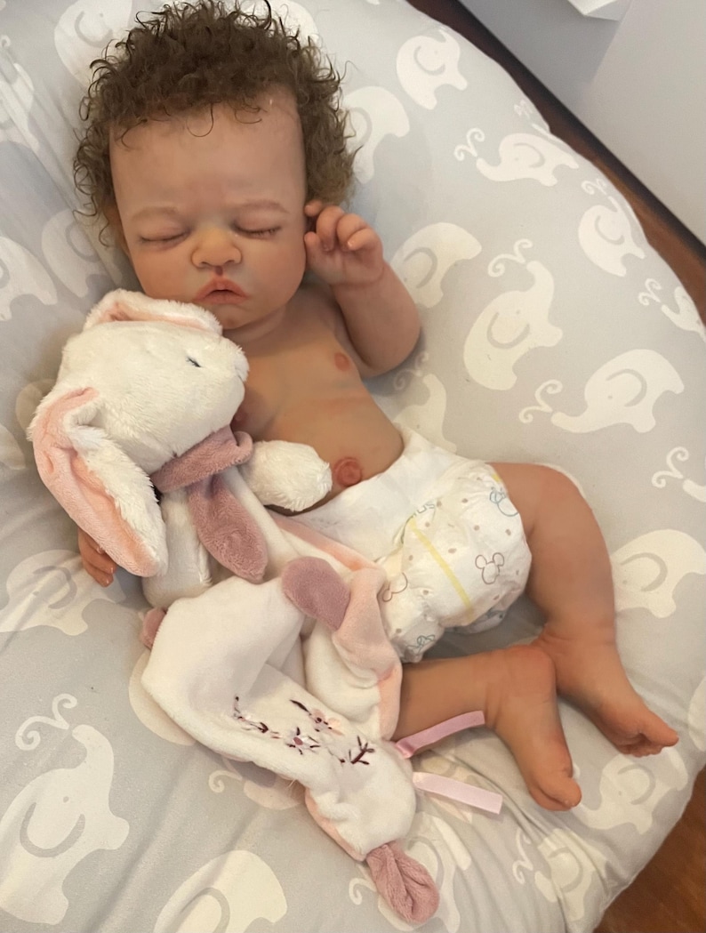 May include: A realistic baby doll with brown curly hair, wearing a nappy, and resting on a grey elephant-patterned pillow. The doll is holding a white and pink plush bunny. The bunny has pink ears and a pink scarf.