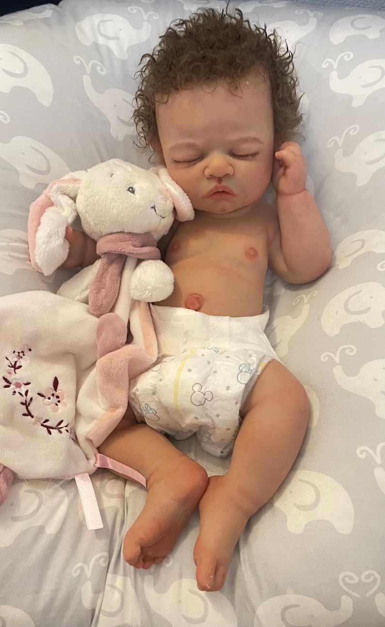 May include: A realistic baby doll with brown curly hair, wearing a nappy, and lying on a grey patterned surface. The doll is holding a white and pink plush bunny and a matching blanket with floral embroidery.