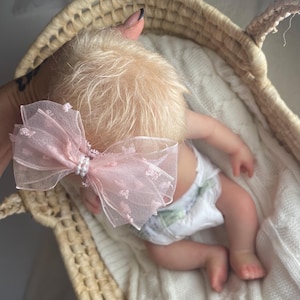 May include: A light pink sheer organza hair bow featuring a textured dotted pattern and a central cluster of small white faux pearls. The bow is attached to a thin band and is positioned on the head of a doll with light blonde hair.