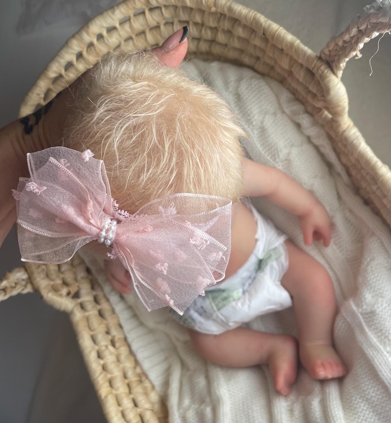 May include: A light pink sheer organza hair bow featuring a textured dotted pattern and a central cluster of small white faux pearls. The bow is attached to a thin band and is positioned on the head of a doll with light blonde hair.