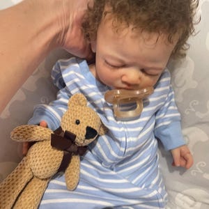 May include: A realistic baby doll with brown curly hair, wearing a blue and white striped onesie and a dummy. A small tan teddy bear with a brown ribbon is held next to the doll.