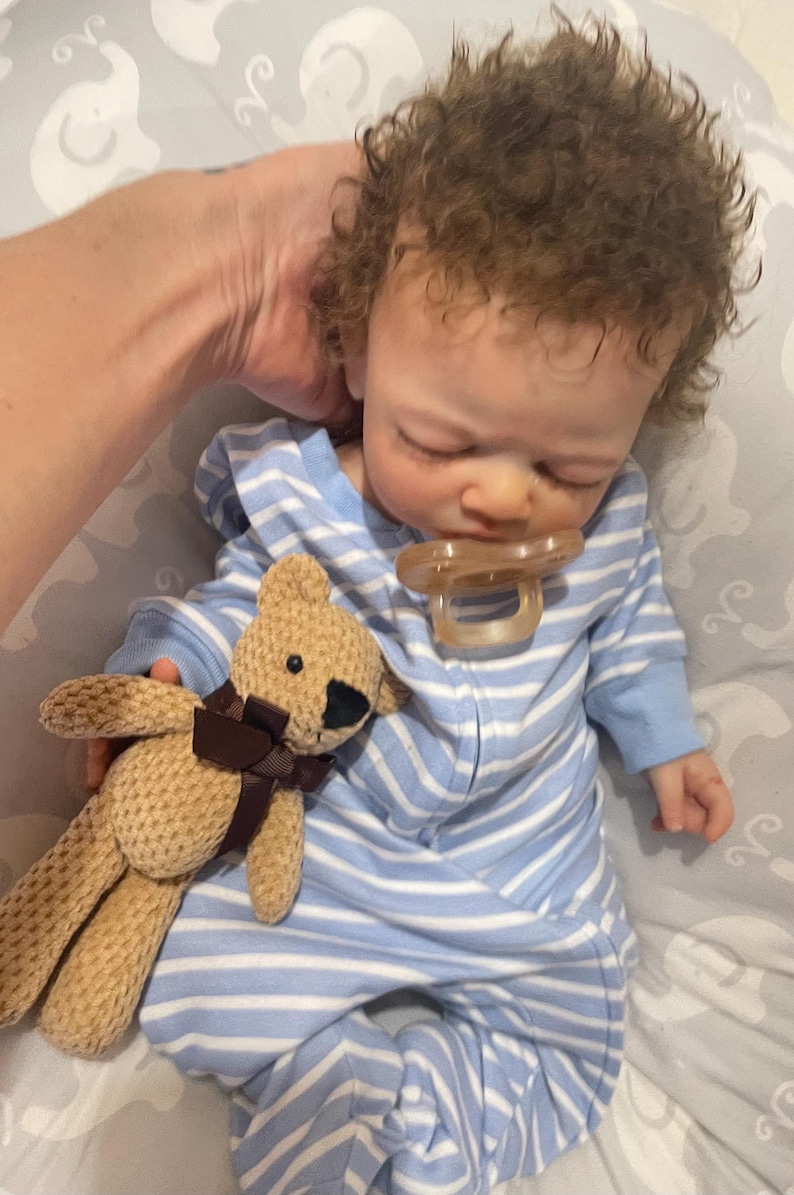 May include: A realistic baby doll with brown curly hair, wearing a blue and white striped onesie and a dummy. A small tan teddy bear with a brown ribbon is held next to the doll.