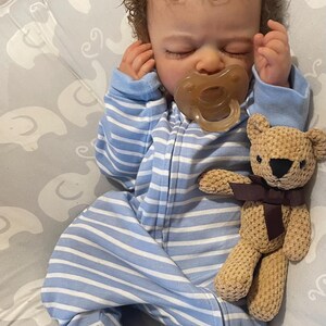 May include: A realistic baby doll dressed in a blue and white striped sleepsuit, with a dummy in its mouth. A small, beige teddy bear with a brown ribbon is next to the doll. The doll has curly brown hair and is lying down.
