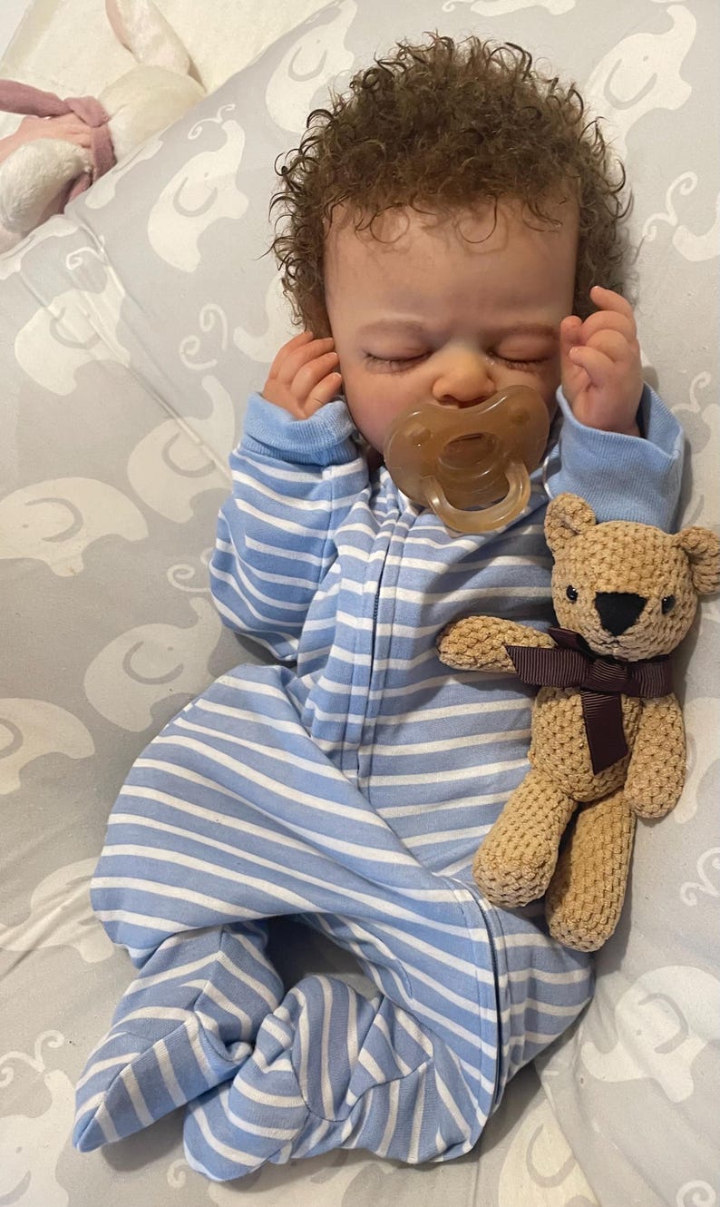 May include: A realistic baby doll dressed in a blue and white striped sleepsuit, with a dummy in its mouth. A small, beige teddy bear with a brown ribbon is next to the doll. The doll has curly brown hair and is lying down.