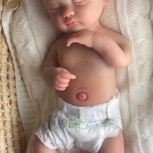 May include: A realistic newborn doll with fair skin, closed eyes, and blonde hair, wearing a white diaper and a light pink lace headband with a bow. The doll is lying on a white knitted blanket inside a woven basket with a wooden button detail.