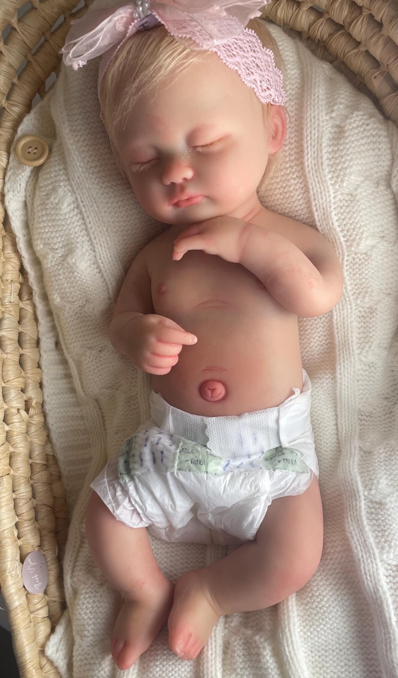 May include: A realistic newborn doll with fair skin, closed eyes, and blonde hair, wearing a white diaper and a light pink lace headband with a bow. The doll is lying on a white knitted blanket inside a woven basket with a wooden button detail.